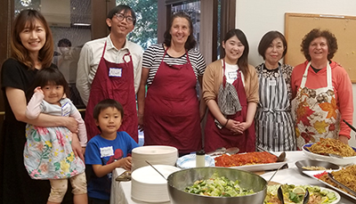 Six smiling internationals and two children behind a table of food they prepared